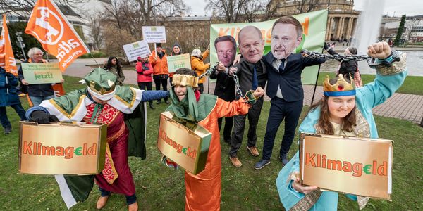Drei als Weise aus dem Morgenland verkleidete Aktivist*innen mit goldenen Klimageldkisten werden mithilfe von Ketten von als Habeck, Lindner und Scholz maskierten Menschen Festgehalten.